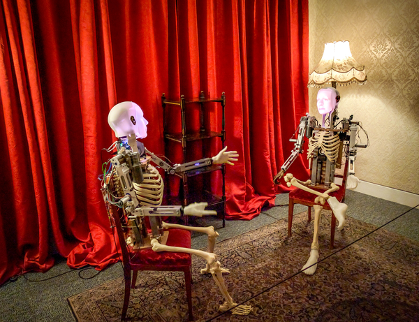 The robot installation in situ at Manchester Art Gallery. The two robots sit on chairs opposite each other, a red curtain behind them. The robots are skeletal, but with faces on their skulls.